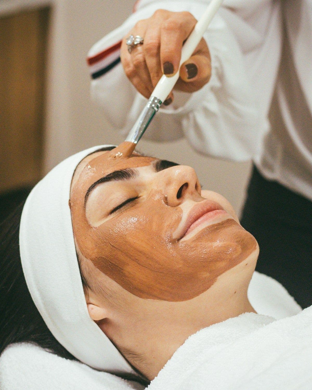 Aesthetician in white applying a treatment mask to a reclining client's face with a brush